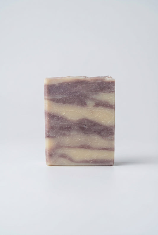 A rectangular bar of marbled soap with cream and dusty lavender-purple swirls, standing on a weathered wooden deck railing with a blurred background of autumn trees.