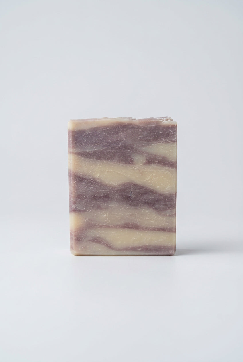 A rectangular bar of marbled soap with cream and dusty lavender-purple swirls, standing on a weathered wooden deck railing with a blurred background of autumn trees.