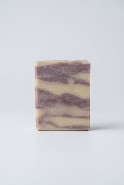 A rectangular bar of marbled soap with cream and dusty lavender-purple swirls, standing on a weathered wooden deck railing with a blurred background of autumn trees.